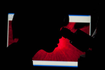 A person looking in a microscope is silhouetted against a red image on a computer screen