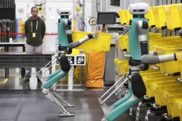 Droid robots stack yellow bins on shelves while a man looks on, arms folded in front of him